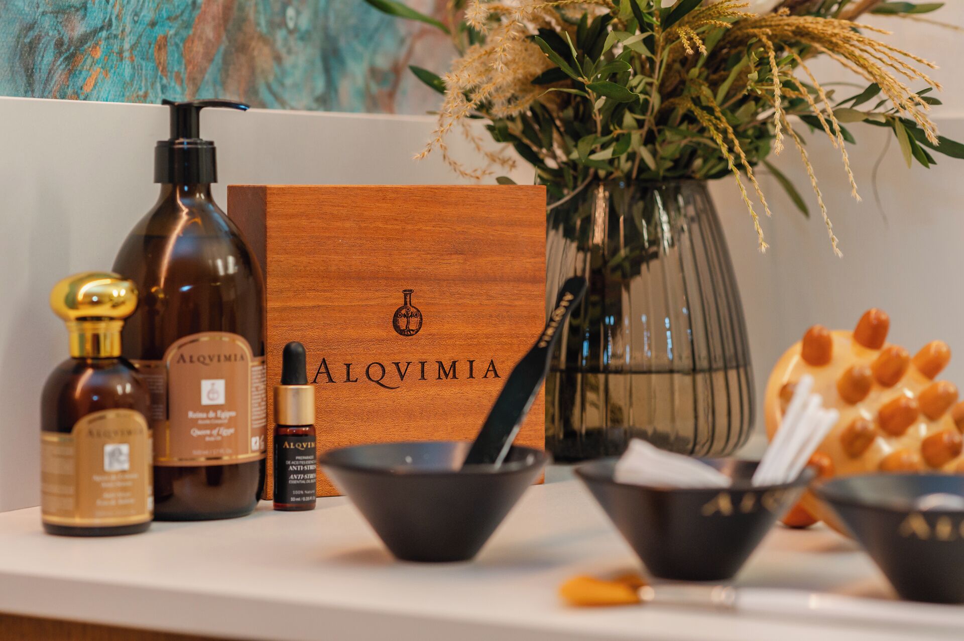 A wooden box with 'ALQVIMIA' branding, a bottle of Alquvimia, a small bottle of essential oil, a bowl, and a vase with dried flowers.