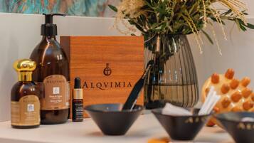 A wooden box with 'ALQVIMIA' branding, a bottle of Alquvimia, a small bottle of essential oil, a bowl, and a vase with dried flowers.