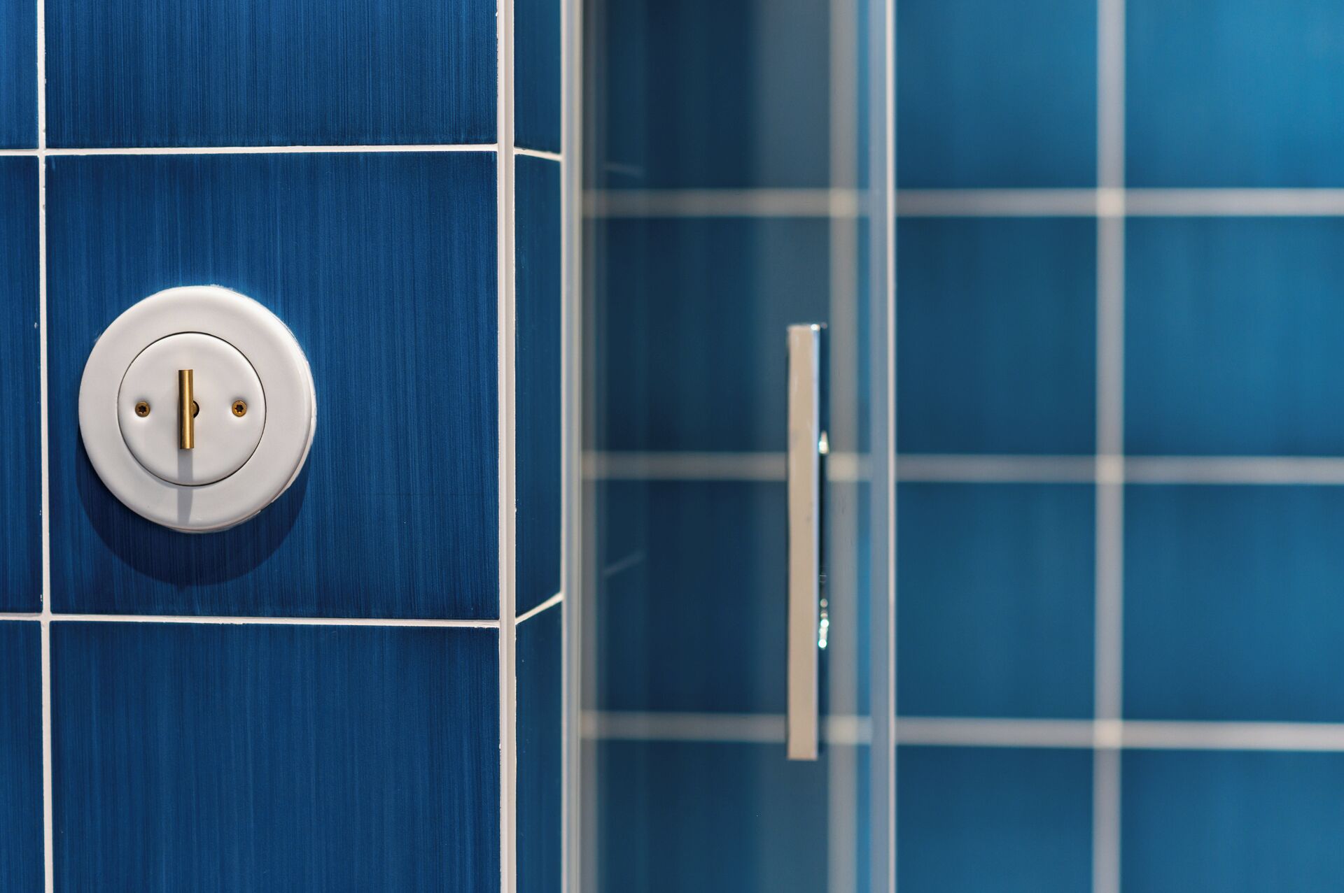 Een badkamer met blauwe betegelde wanden, een witte deur en een ronde lichtschakelaar.