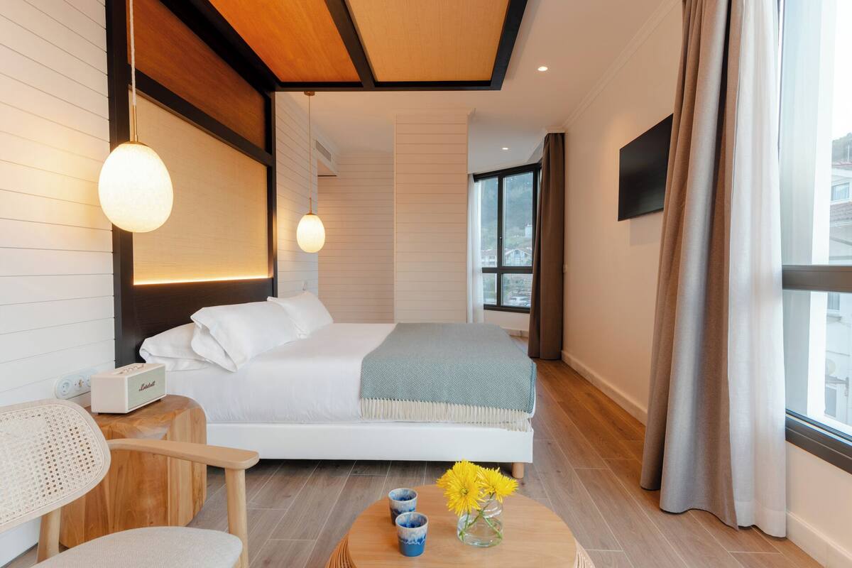 Ein modernes Hotelzimmer mit einem großen Bett, einem Holzkopfstück, einem Nachttisch mit Lampe, Blick auf die Stadt durch das Fenster und einem kleinen Tisch mit Blumen in einer Vase.