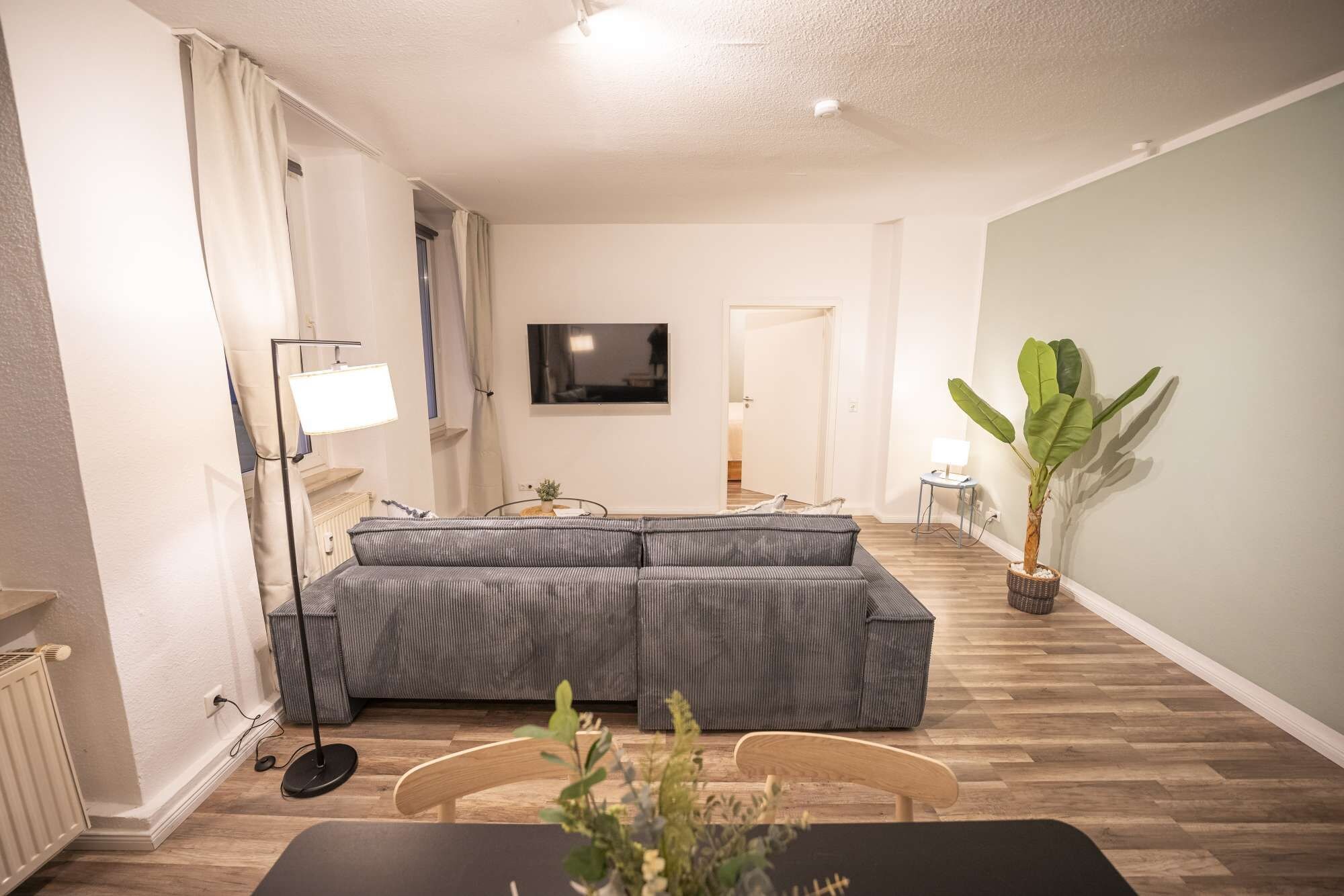 Un salon comprenant un canapé, un téléviseur fixé au mur, une plante en pot et un coin repas avec une table et des chaises.