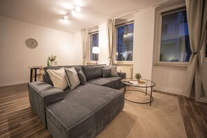 Een woonkamer met een grijze bank, een klein rond salontafeltje en een klok aan de muur.