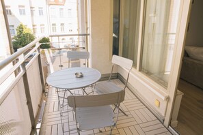 Un balcon avec une table et deux chaises, une fenêtre avec des rideaux et une vue sur des immeubles.