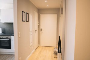 Un couloir étroit, avec une porte blanche au fond, un coin cuisine avec une cuisinière et des placards sur la gauche, et un porte-chaussures avec des chaussures sur la droite.