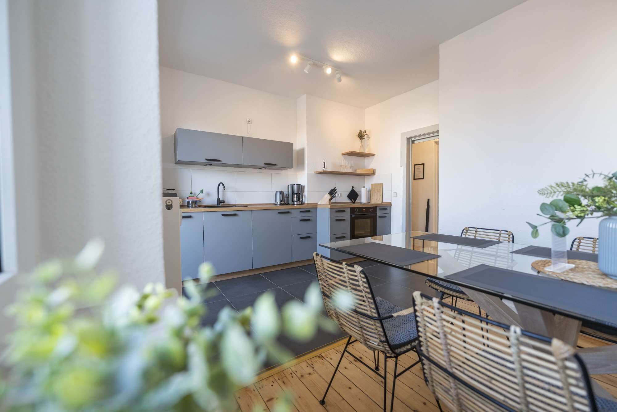 A modern kitchen with grey cabinets, a black countertop, and a dining area with a glass table and wicker chairs.