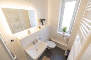 A modern bathroom with a white sink, toilet, and a window with a plant. - RR | Central Apartment 80 m² -2 | Balcony | Washer (Magdeburg)