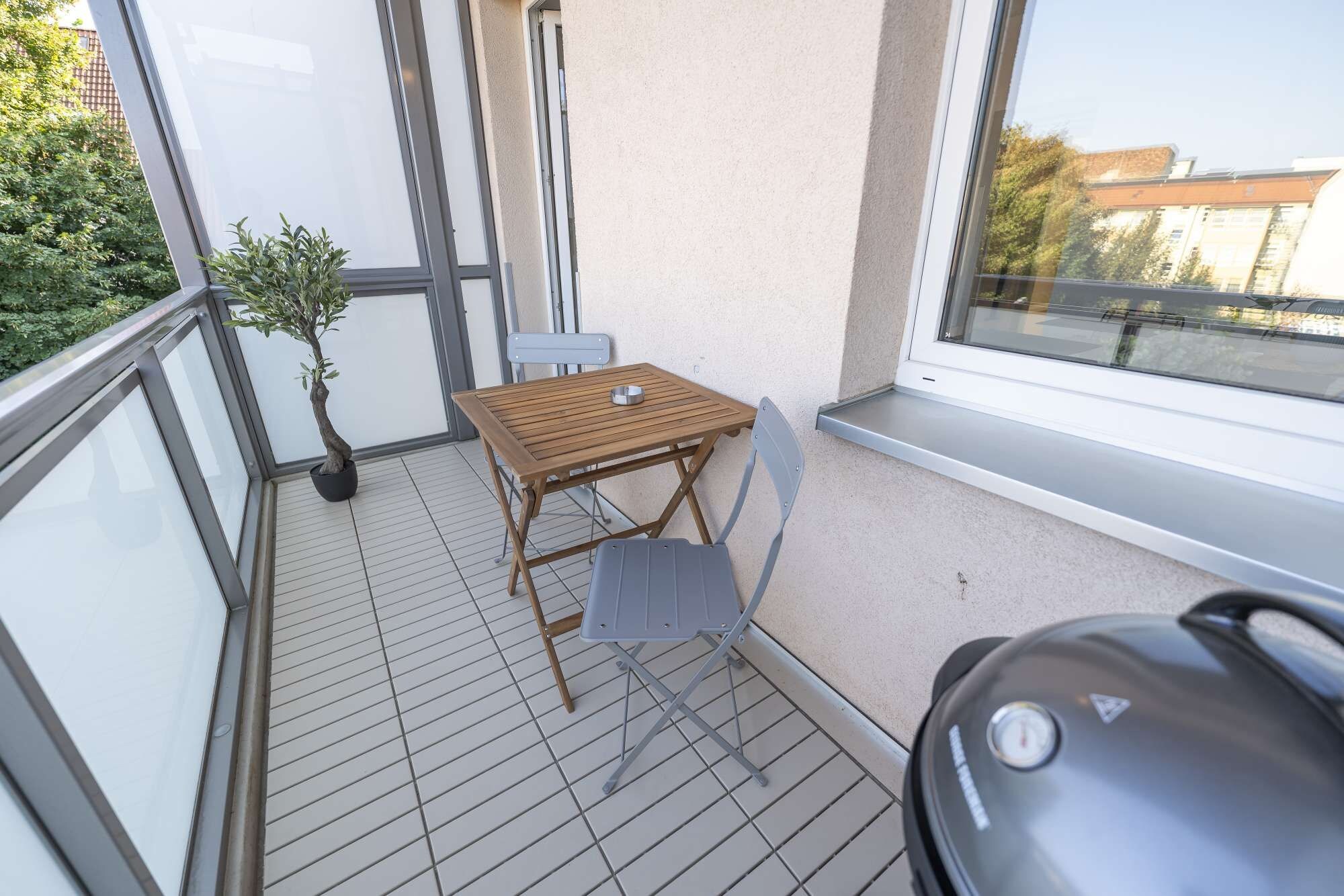 A balcony with a wooden table and chairs, a potted plant, and a barbecue grill.