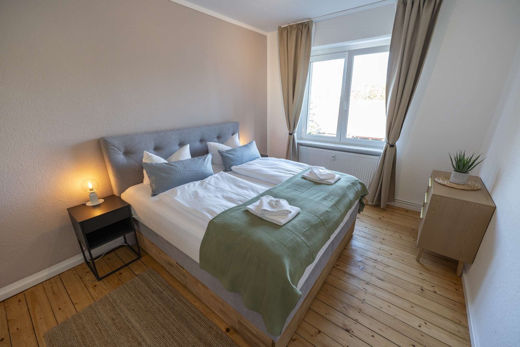 A bedroom with a bed, bedside table, window, curtains, and a small plant.