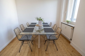 Een kamer met een tafel met glazen blad, omringd door rieten stoelen, een potplant en een raam met uitzicht.