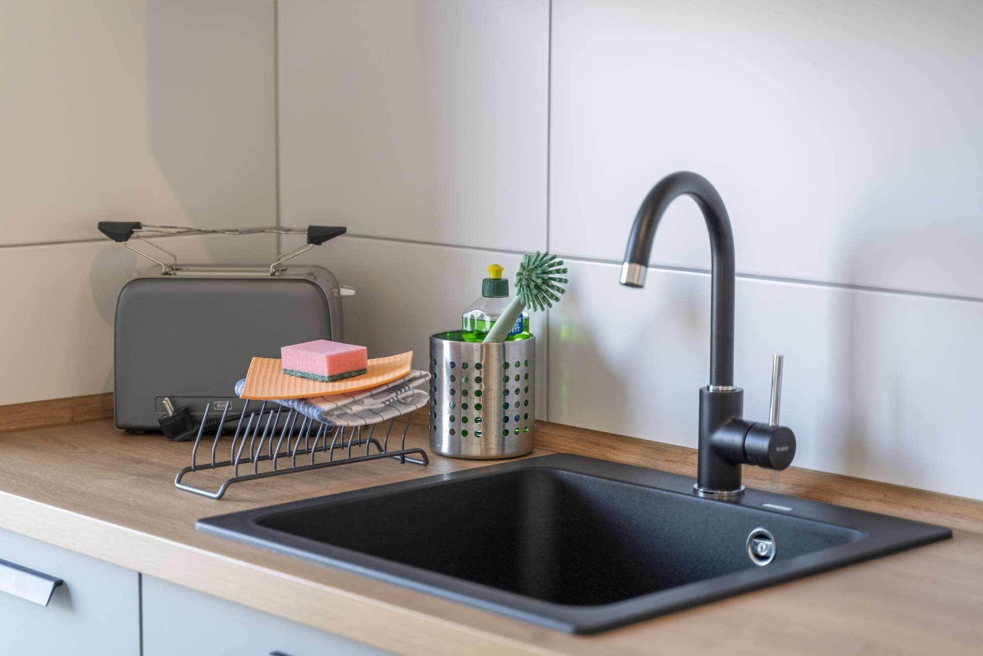 Een moderne keuken met een zwarte spoelbak, een klein draadrooster, een groene fles en een roze spons.