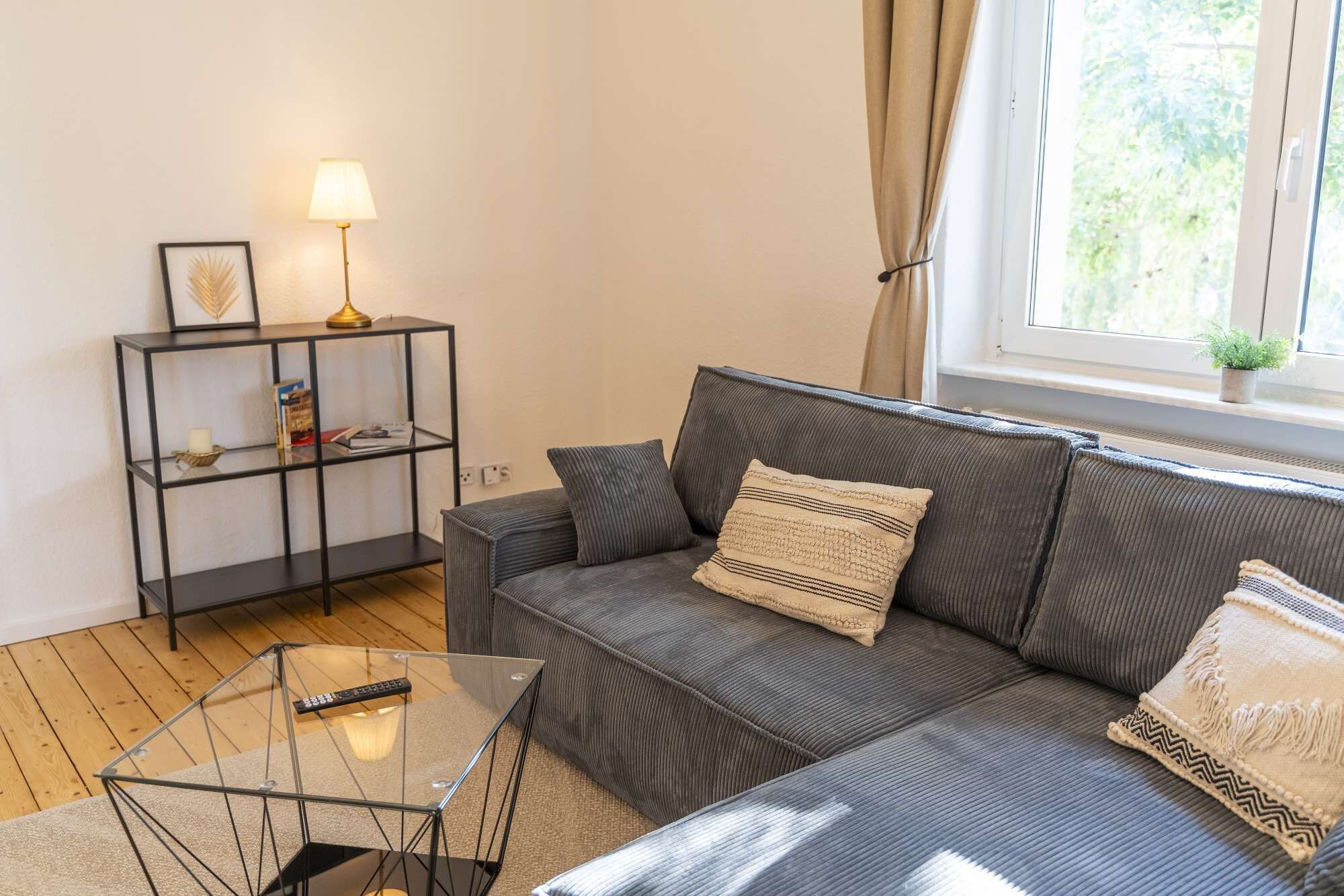 Een woonkamer met een grijze bank, een glazen salontafel, een zwart metalen bijzettafeltje met een lamp, en een ingelijste foto aan de muur.