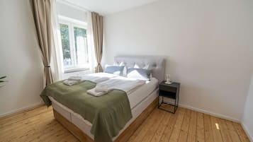 Een slaapkamer met een bed, een nachtkastje, een raam met gordijnen en een houten vloer.
