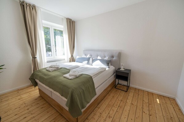 A bedroom with a bed, a bedside table, a window with curtains, and a wooden floor.