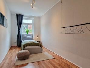 Une chambre à coucher comprenant un lit, une table de chevet, une lampe, un portant à vêtements et une fenêtre donnant sur des arbres.