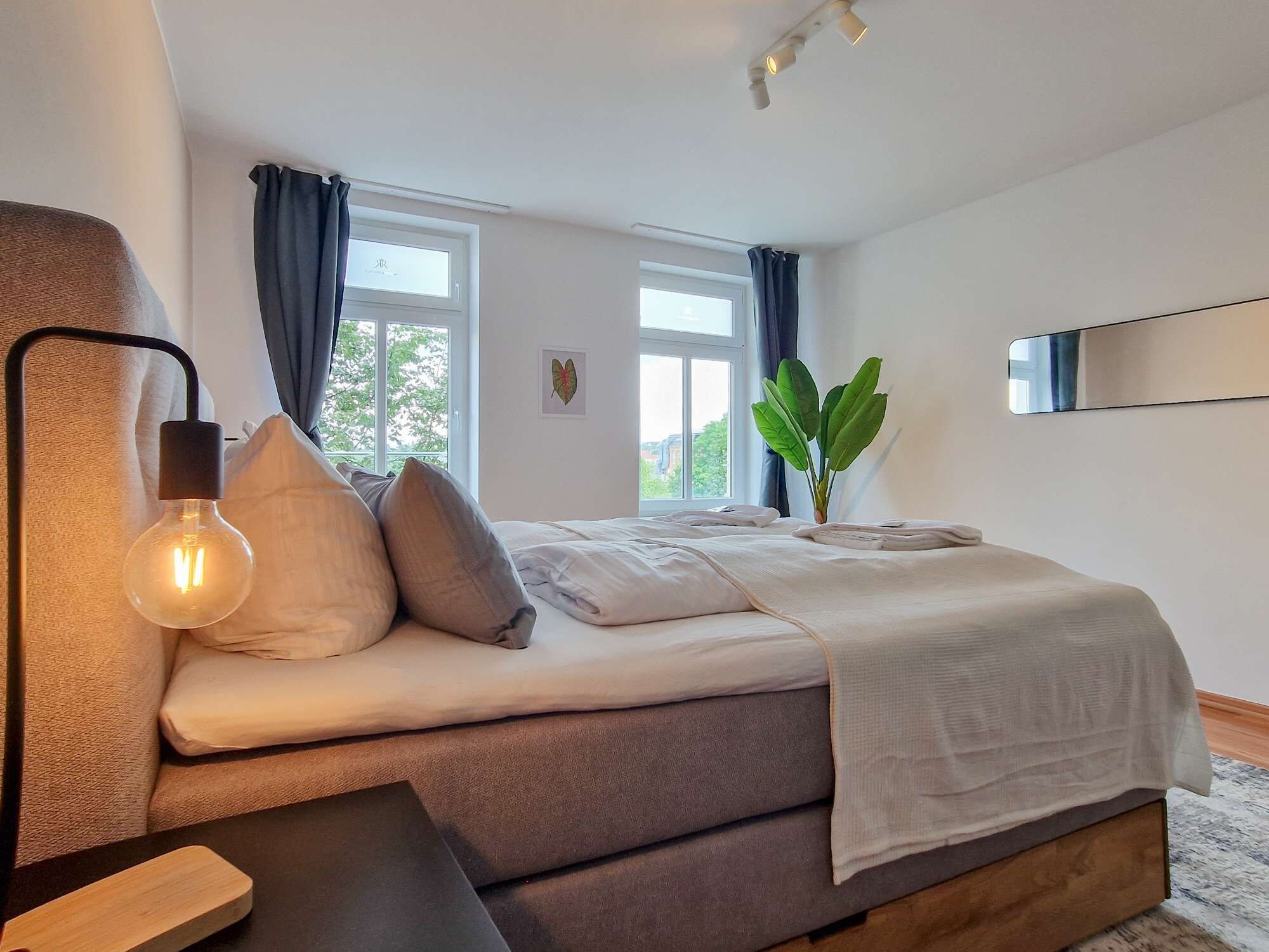 A modern bedroom with a bed, a bedside lamp, a plant, and a window.