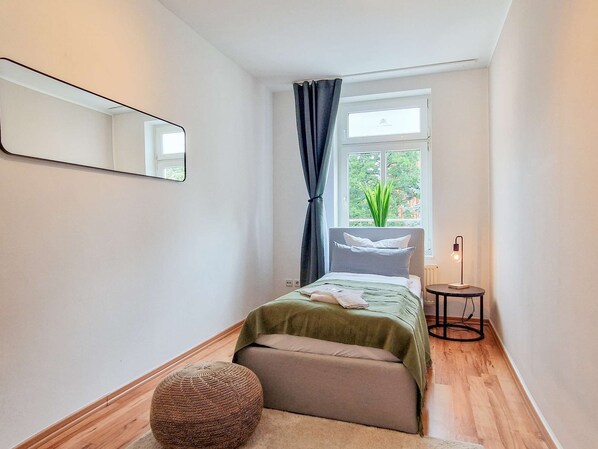 A small, well-lit bedroom with a bed, a bedside table, a lamp, a window with curtains, and a mirror on the wall.
