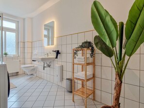 Une salle de bain moderne avec des toilettes, un lavabo et un miroir. On y trouve une plante et une étagère avec des serviettes.
