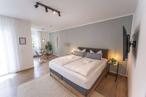 Een moderne slaapkamer met een tweepersoonsbed, nachtkastjes, een televisie en een zithoek met een tafel en stoelen.