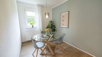 Een eetgedeelte met een glazen tafel, twee stoelen, een potplant en een lijst met een afbeelding aan de muur.