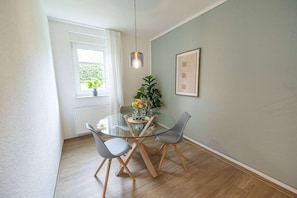 Een eetgedeelte met een glazen tafel, twee stoelen, een potplant en een lijst met een afbeelding aan de muur.
