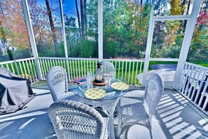 Une terrasse couverte, aménagée avec des meubles en osier, une table en verre et offrant une vue sur les arbres et la pelouse.