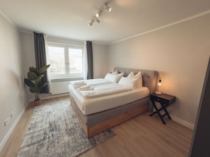 A modern bedroom with a large bed, a bedside table with a lamp, a window with curtains, and a potted plant.
