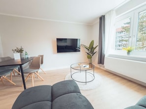 Ein modernes Wohnzimmer mit Fernseher, Esstisch mit Stühlen, rundem Couchtisch und großem Fenster mit Blick auf Gebäude.