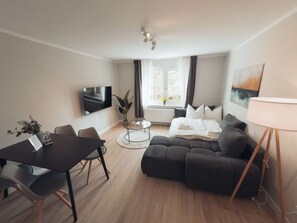 A modern living room with a dark sofa, a dining table with chairs, a flat-screen TV mounted on the wall, and a lamp on a side table.