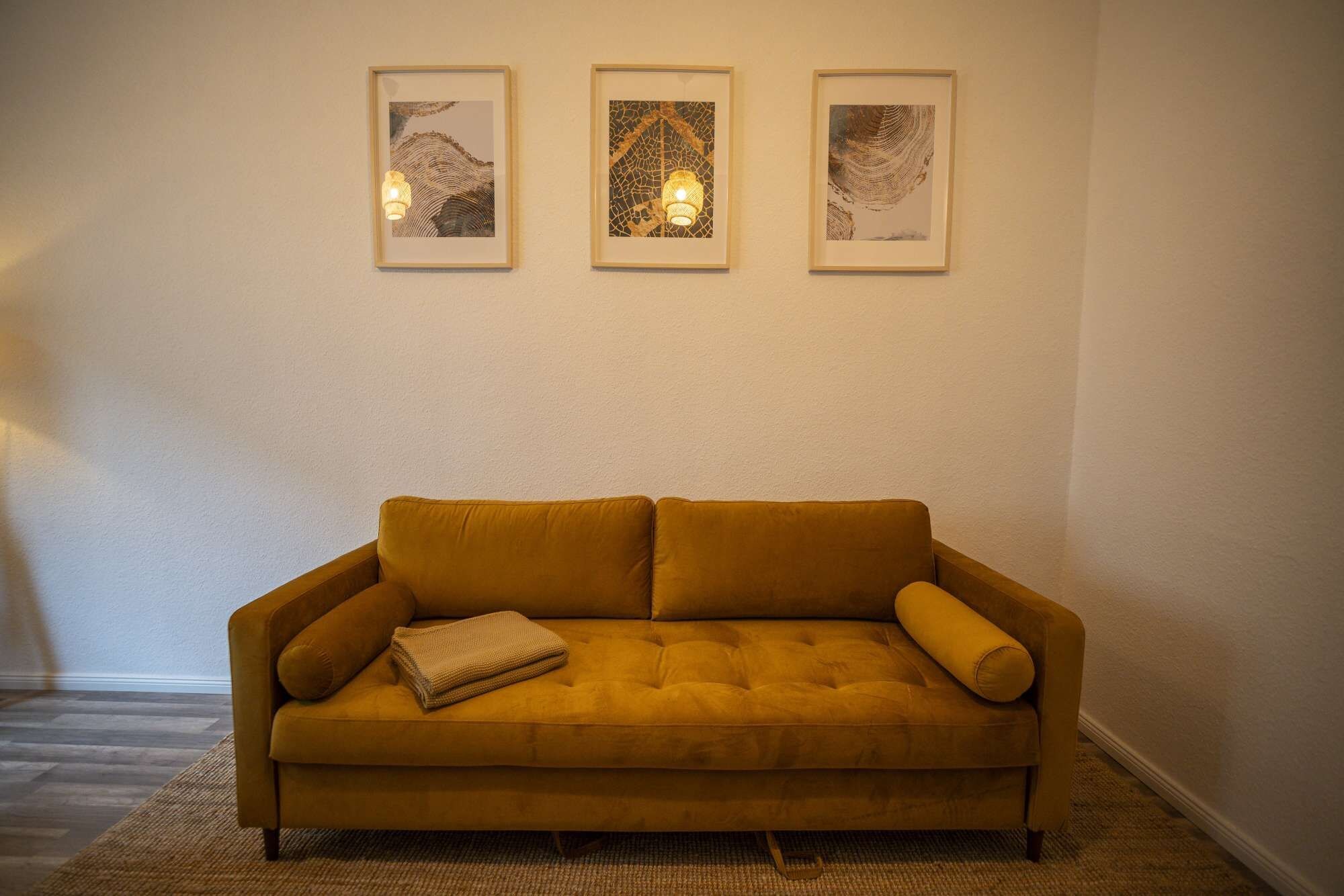 A mustard-colored sofa with a folded blanket, three framed artworks on the wall, and a lamp.