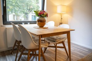A dining area with a wooden table, white chairs, a vase with flowers, a lamp, and a view of trees through a window.