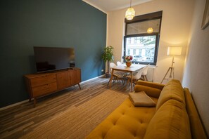 A living room with a sofa, a television on a wooden cabinet, a dining table with chairs, a potted plant, and a window with blinds.
