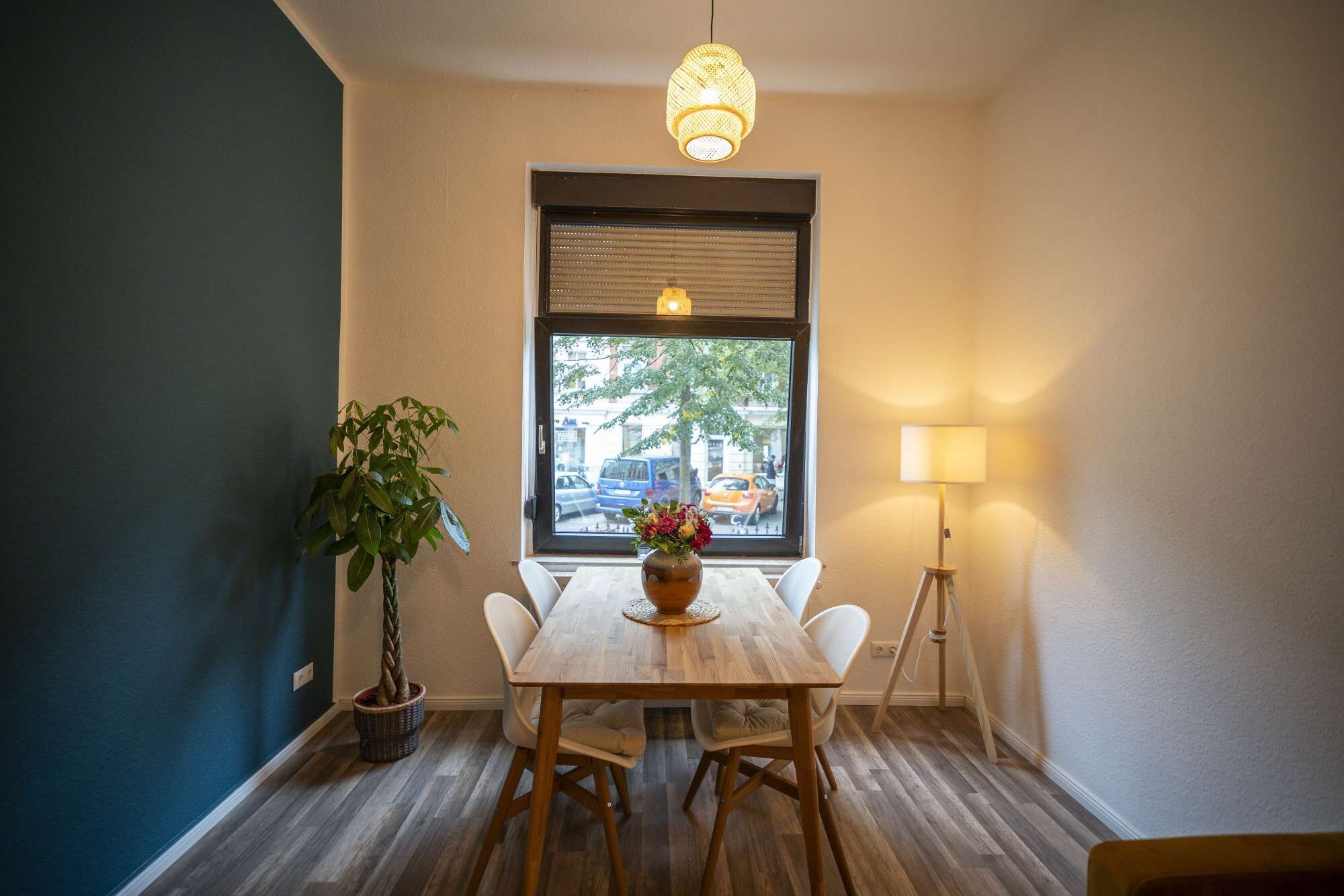 Ein Essbereich mit Tisch, Stühlen, einer Zimmerpflanze, einem Fenster mit Jalousien und einer Stehlampe.