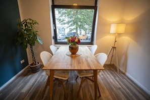 Un comedor con una mesa de madera, sillas blancas, una planta en maceta y un ventanal con vistas a la calle.
