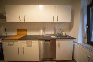 A compact kitchen with white cabinets, a stainless steel dishwasher, and a wooden cutting board. - RR | Spacious Apt_1-70 m² | Parking | Central (Magdeburg)