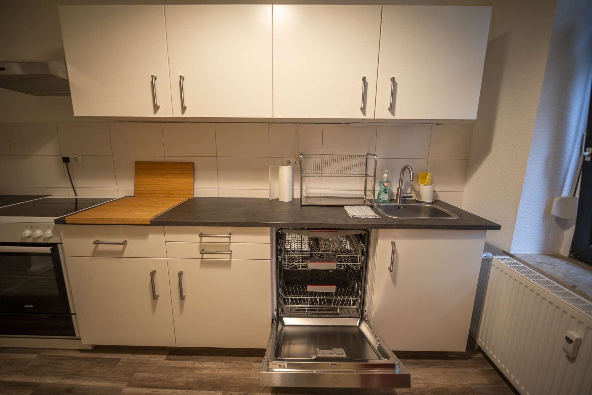 Una cocina compacta con armarios blancos, una tabla de cortar de madera, un lavavajillas empotrado y una estufa.
