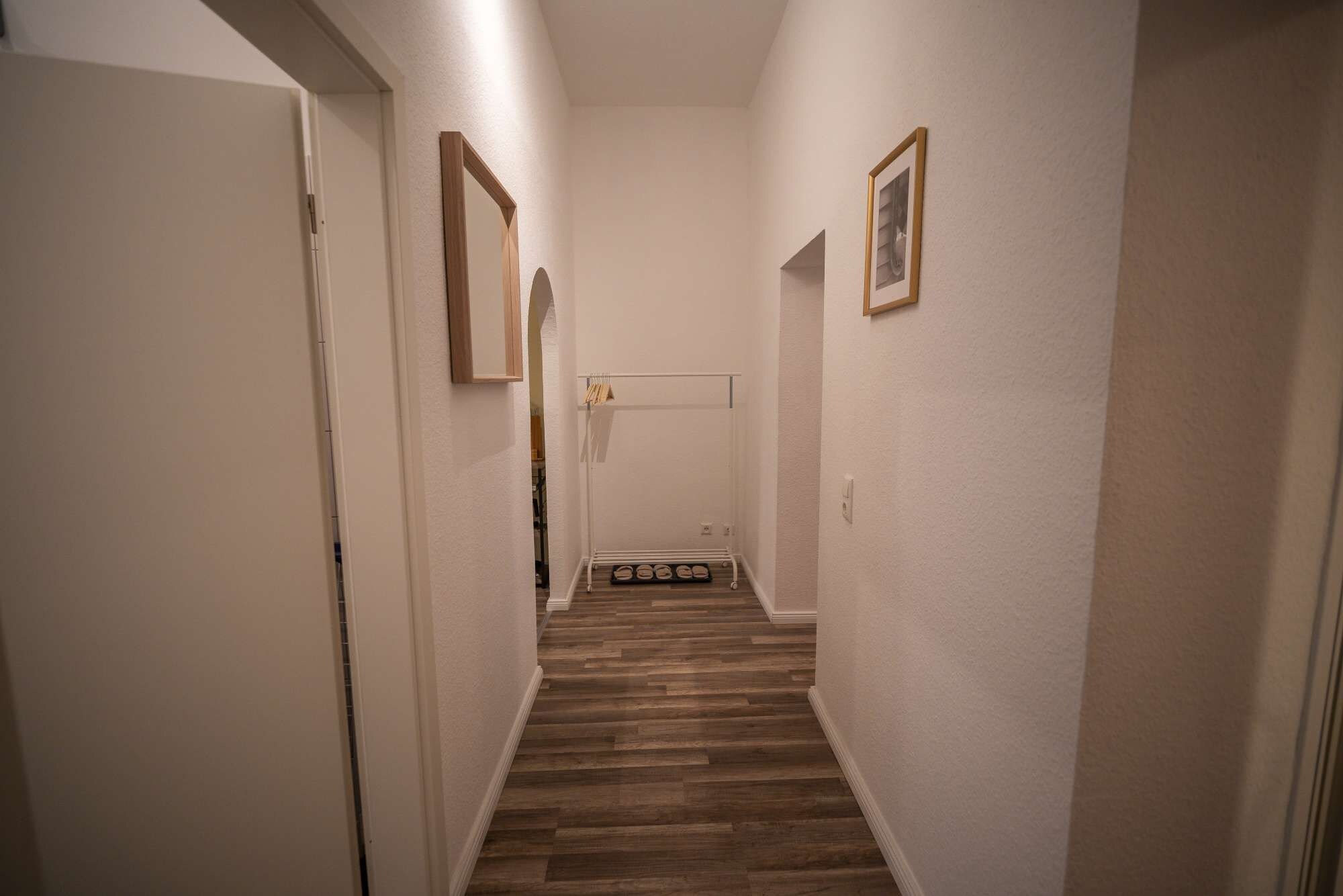 Un pasillo estrecho con suelo de madera, paredes blancas y una puerta al final.
