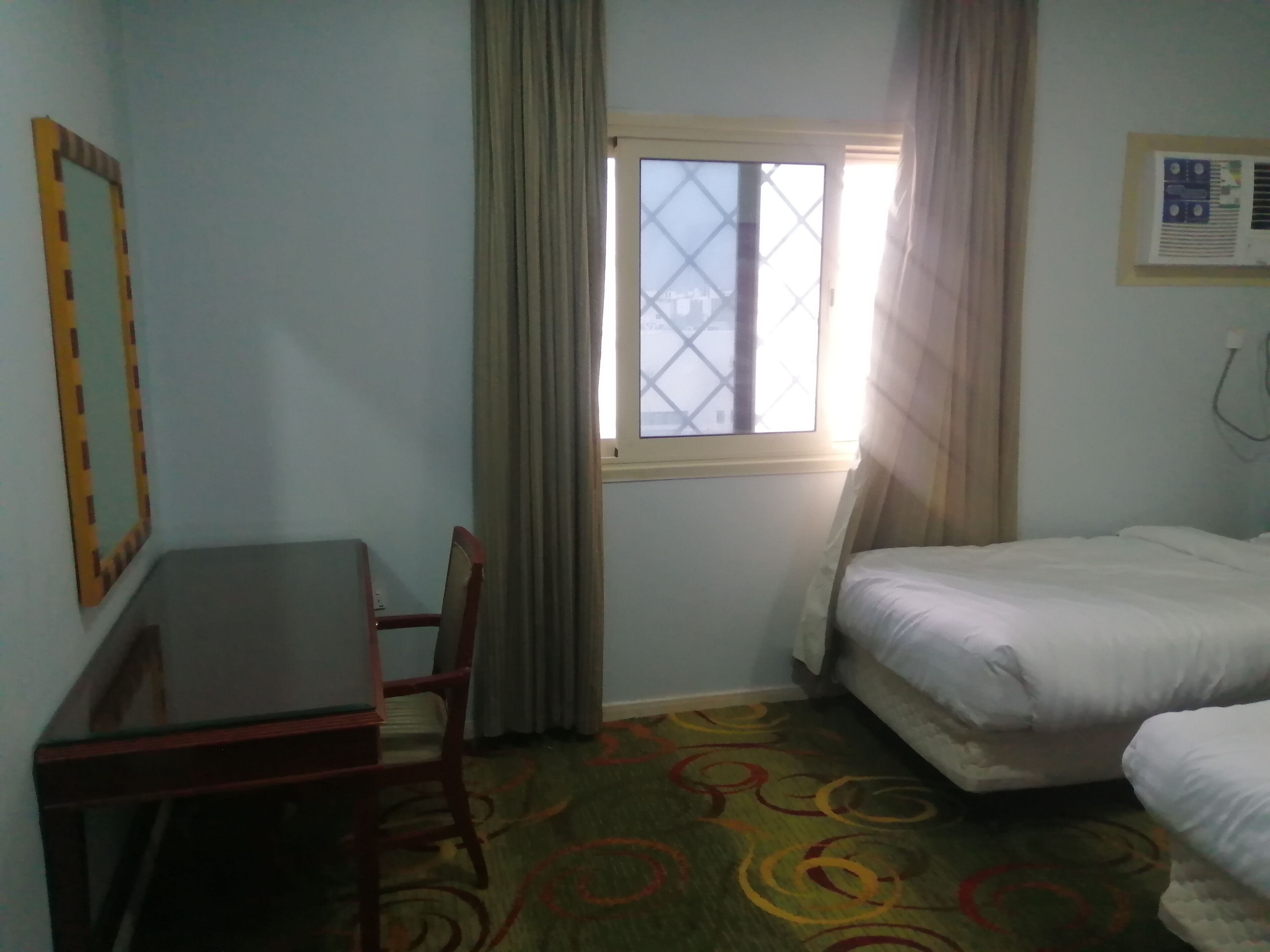 A small hotel room with a single bed, a desk, and a chair. There is a window with curtains, and a wall-mounted air conditioning unit.
