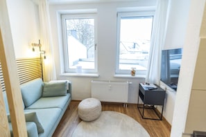 Ein modernes Hotelzimmer mit einer Couch, einem runden Hocker, einer Heizung, einem Schreibtisch mit Fernseher und zwei mit Vorhängen versehenen Fenstern.