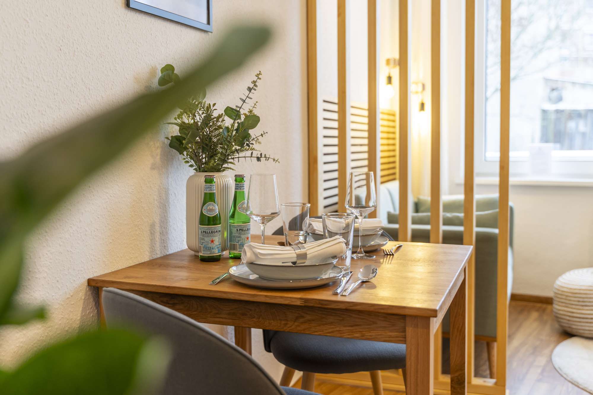 Une table à manger dressée avec des assiettes, des verres et des couverts, un vase avec des plantes vertes et deux bouteilles d’eau Pellegrino.