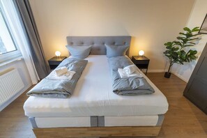 Una cama doble con cabecera gris, dos mesitas de noche con lámparas, una planta en maceta y un radiador.