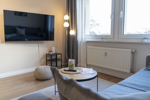 Un salón moderno con un televisor de pantalla plana montado en la pared, una mesa de centro redonda con un control remoto, una mesita auxiliar con una planta y un gran ventanal que proporciona luz natural.
