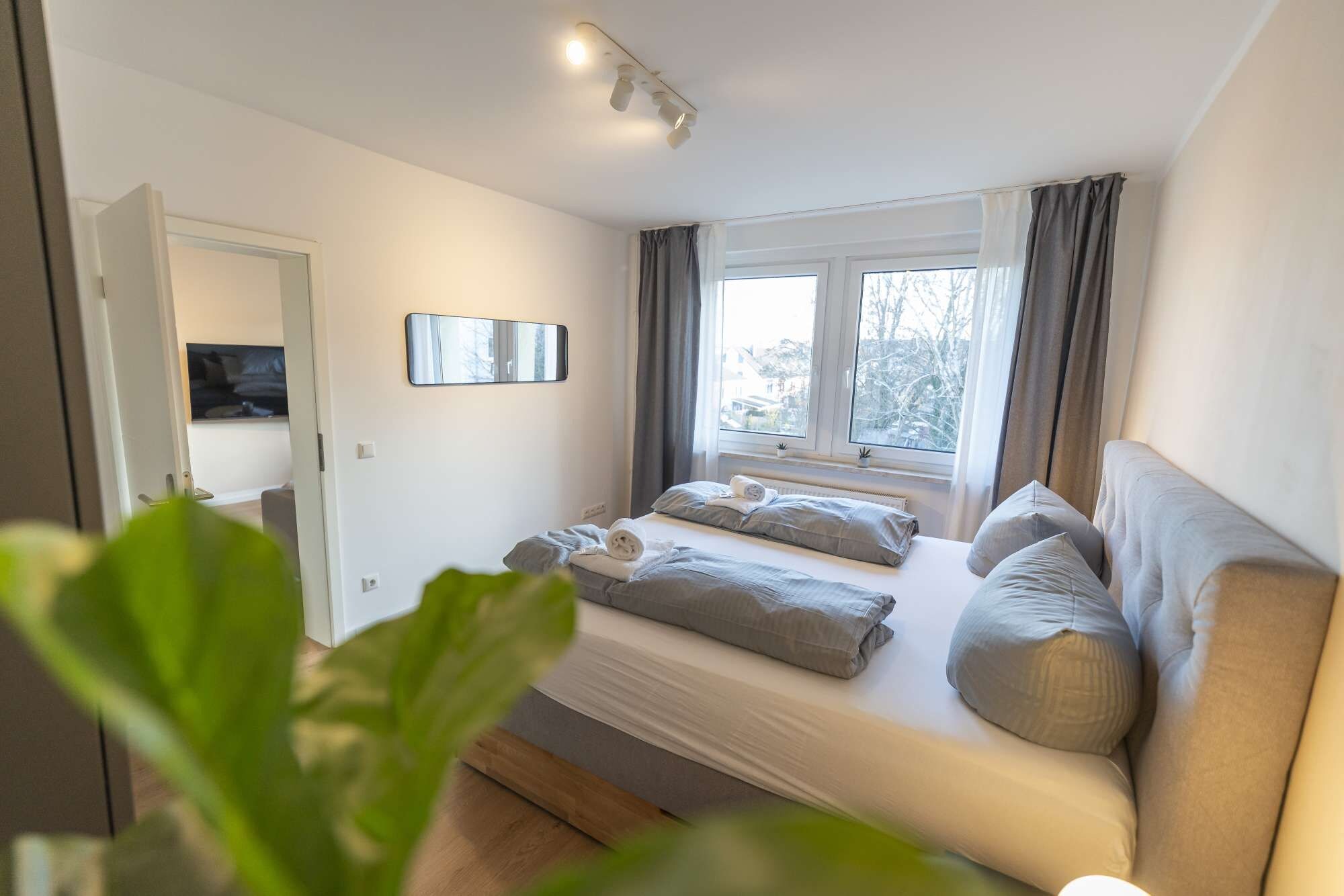 Un dormitorio con cama, mesita de noche, espejo y ventana con cortinas.