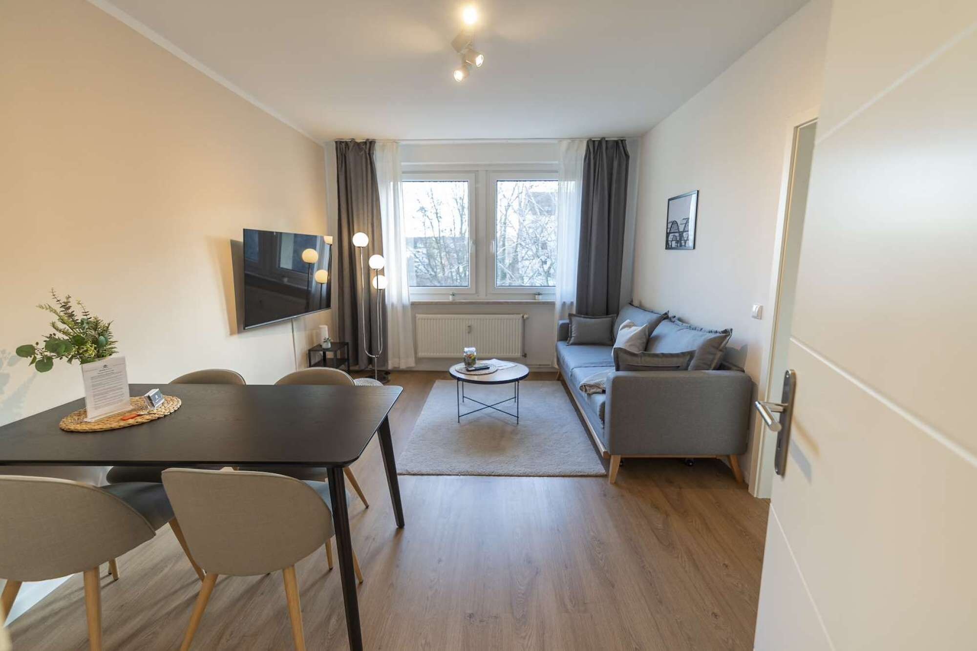 Ein modernes Wohnzimmer mit einer Couch, einem Esstisch mit Stühlen, einem Fernseher und einem Fenster mit Vorhängen.