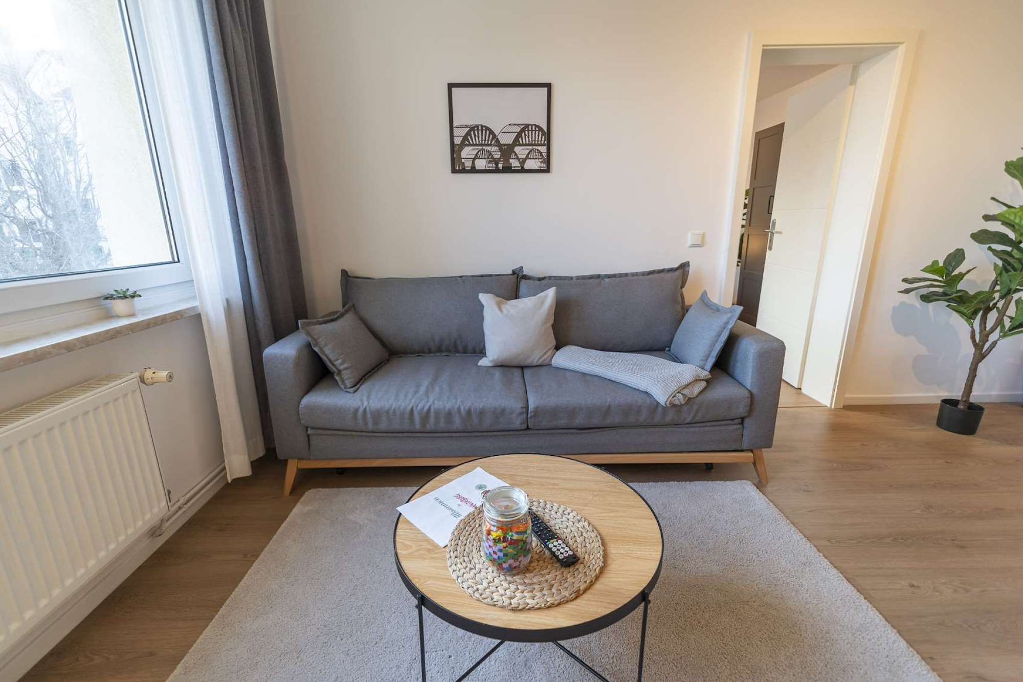 Ein Wohnzimmer mit einem grauen Sofa, einem kleinen runden Couchtisch mit Fernbedienung und einer Süßigkeitsglase, einem gerahmten Bild an der Wand und einer Zimmerpflanze.