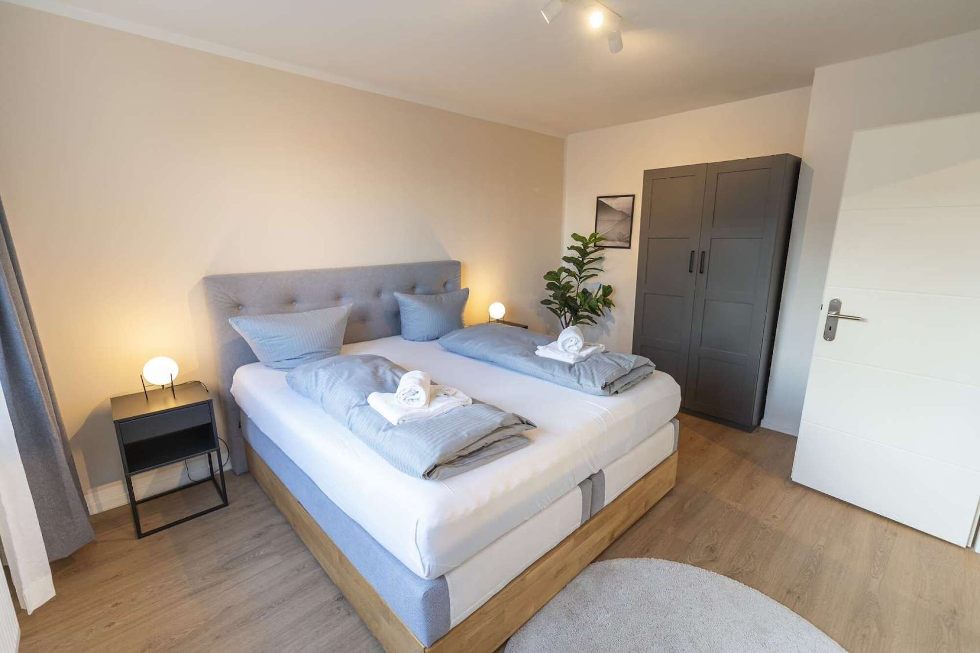 Una cama doble con cabecera gris, dos mesitas de noche con lámparas, un armario gris y una puerta blanca.