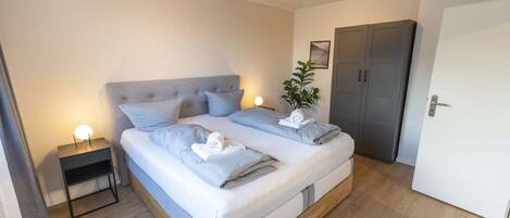 A double bed with a grey headboard, two bedside tables with lamps, a grey wardrobe, and a white door.