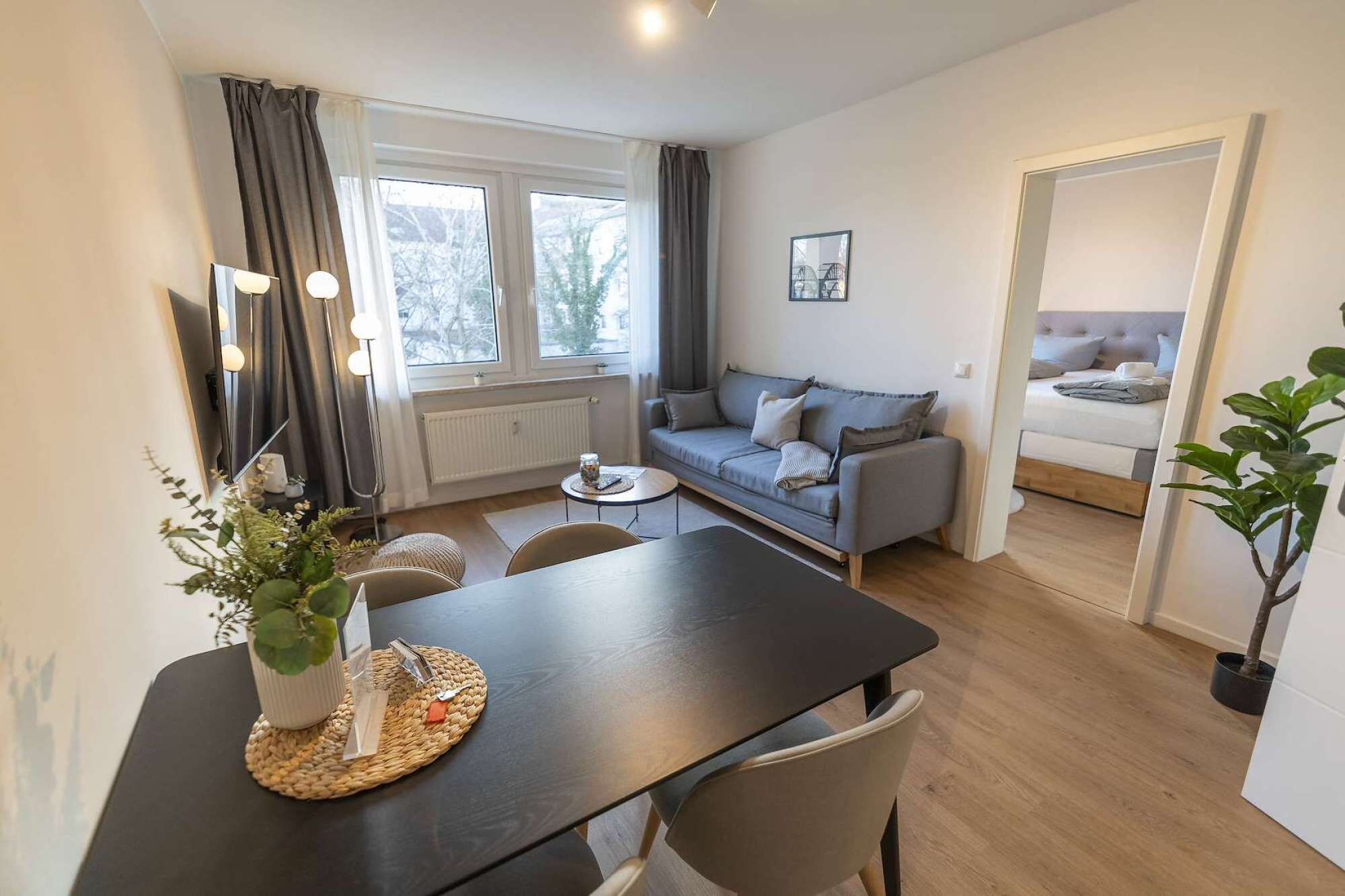 Ein modernes Wohnzimmer mit einer Couch, einem Esstisch mit Stühlen und Blick auf ein Schlafzimmer durch eine offene Tür.