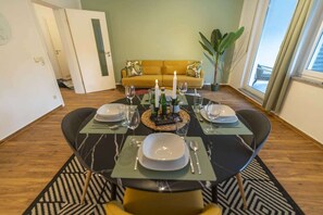 Mesa de comedor para cuatro personas con diseño moderno, un sofá al fondo y un amplio ventanal con cortinas.
