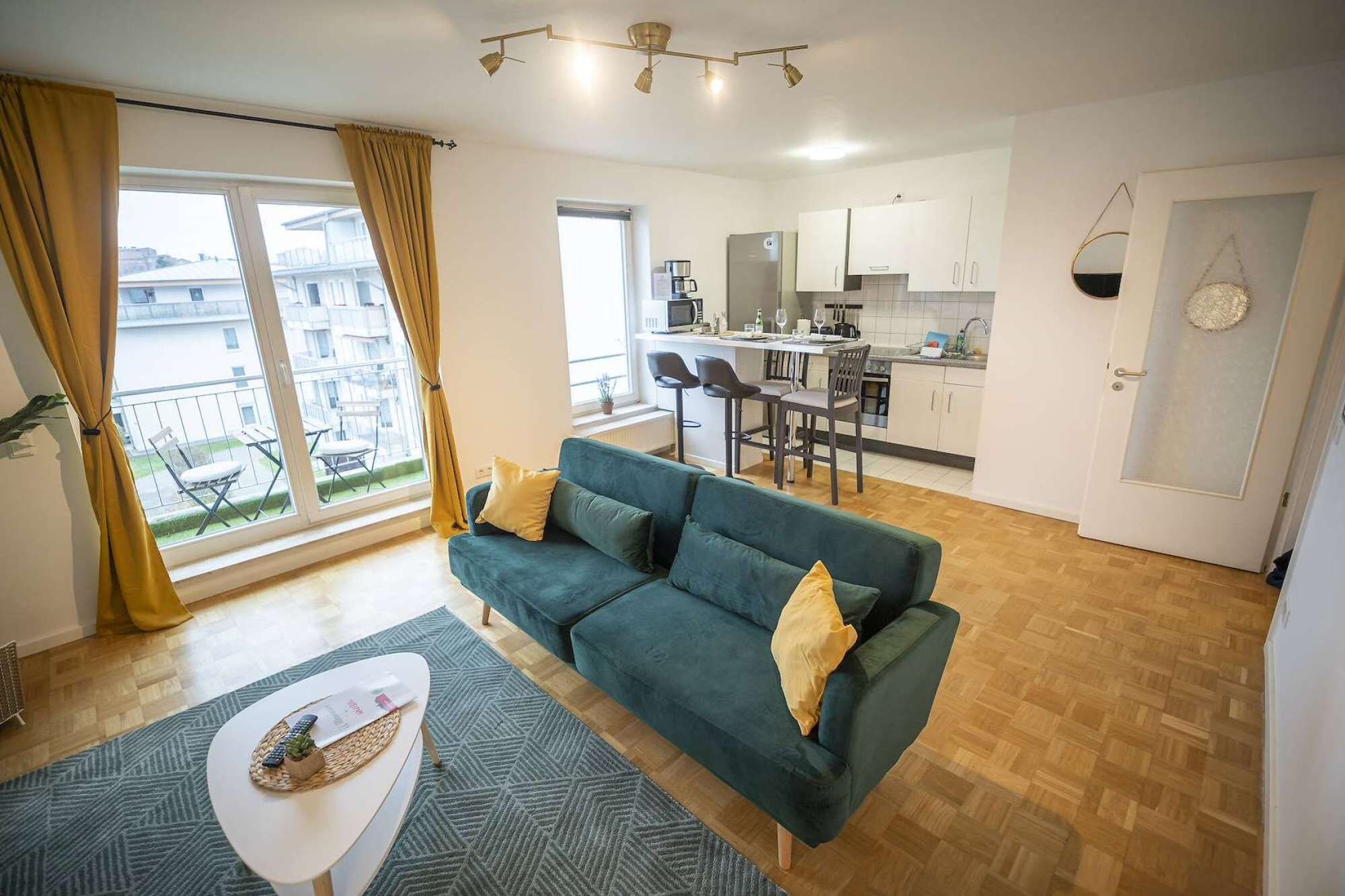 A modern living room with a green sofa, a dining area with a kitchen in the background, and a balcony with yellow curtains.