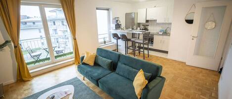 A modern living room with a green sofa, a dining area with a kitchen in the background, and a balcony with yellow curtains.